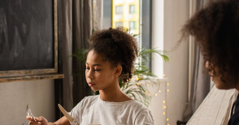 Etiquette - Serious African American girl with unrecognizable mother sitting at wooden table with plate and silverware in hands learning table manners