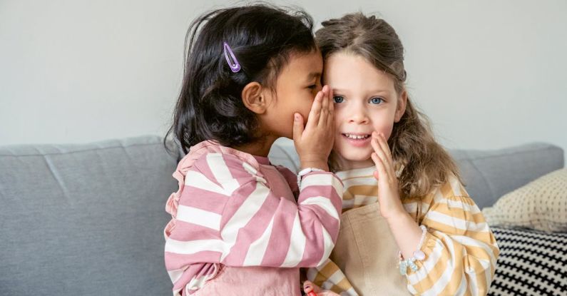 Confidentiality - Ethnic girl sitting on sofa close to smiling best friend and whispering secrets on ear